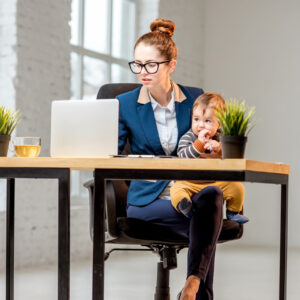 Young multitasking businessmam dressed in the suit working with laptop and documents sitting with her baby son at the office